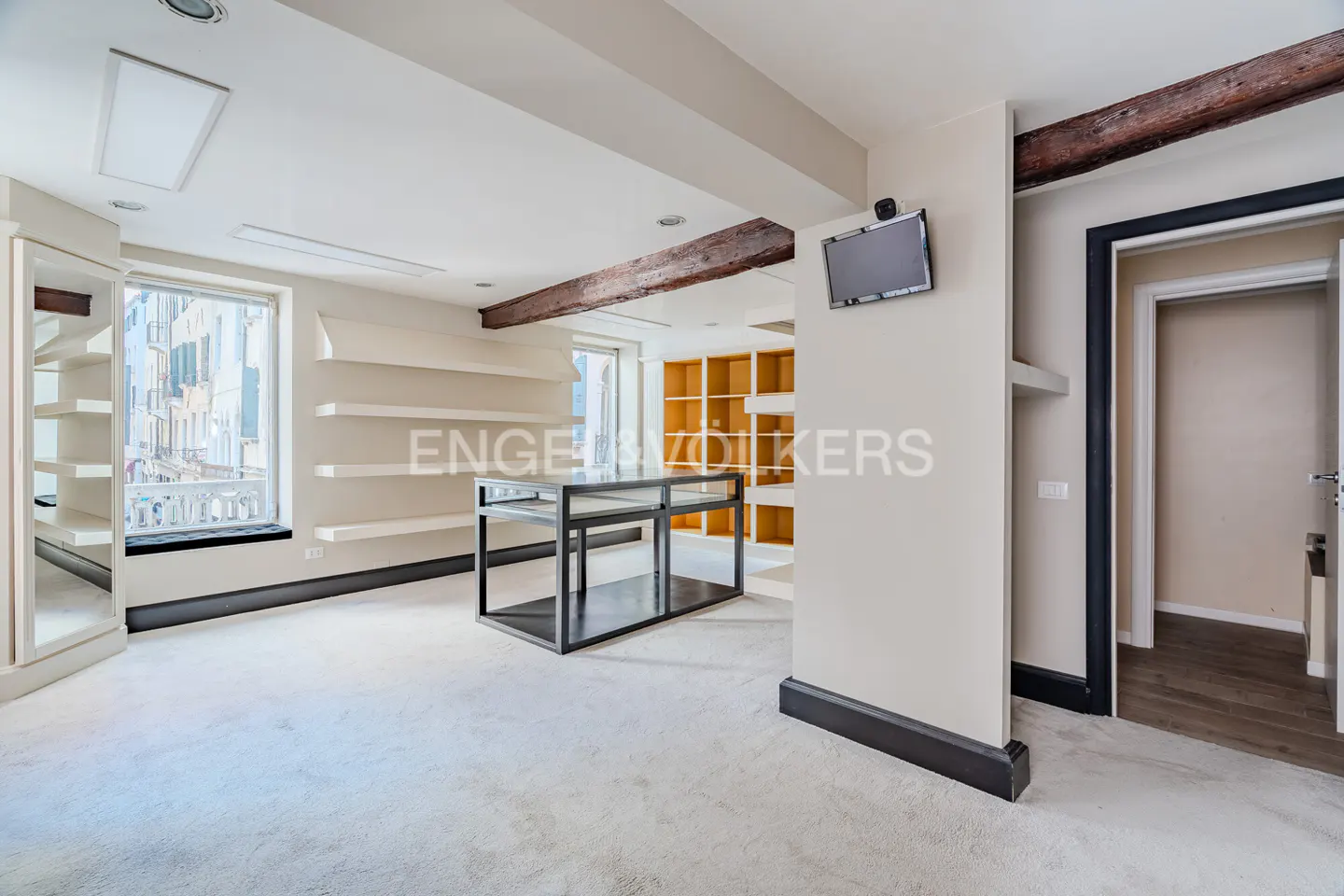 Bright room with white walls, wooden beams, and shelves. A black metal table sits in the center, with a TV mounted on the wall.
