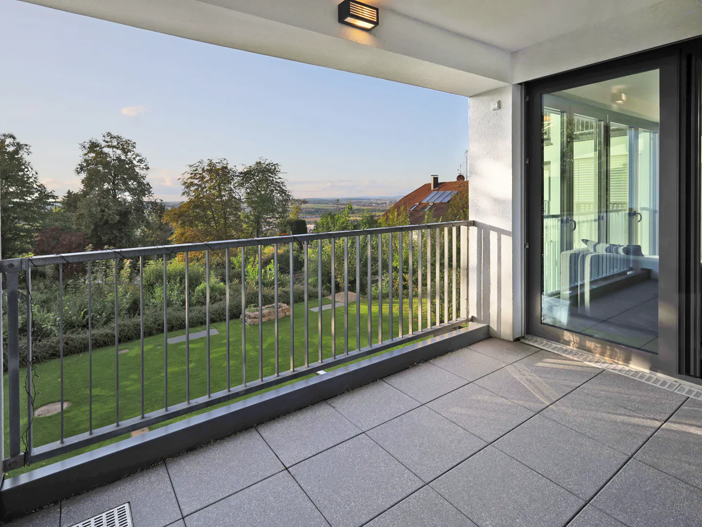 Balcony with gray tile floor, metal railing, and sliding glass door. View of green lawn, trees, and distant buildings.
