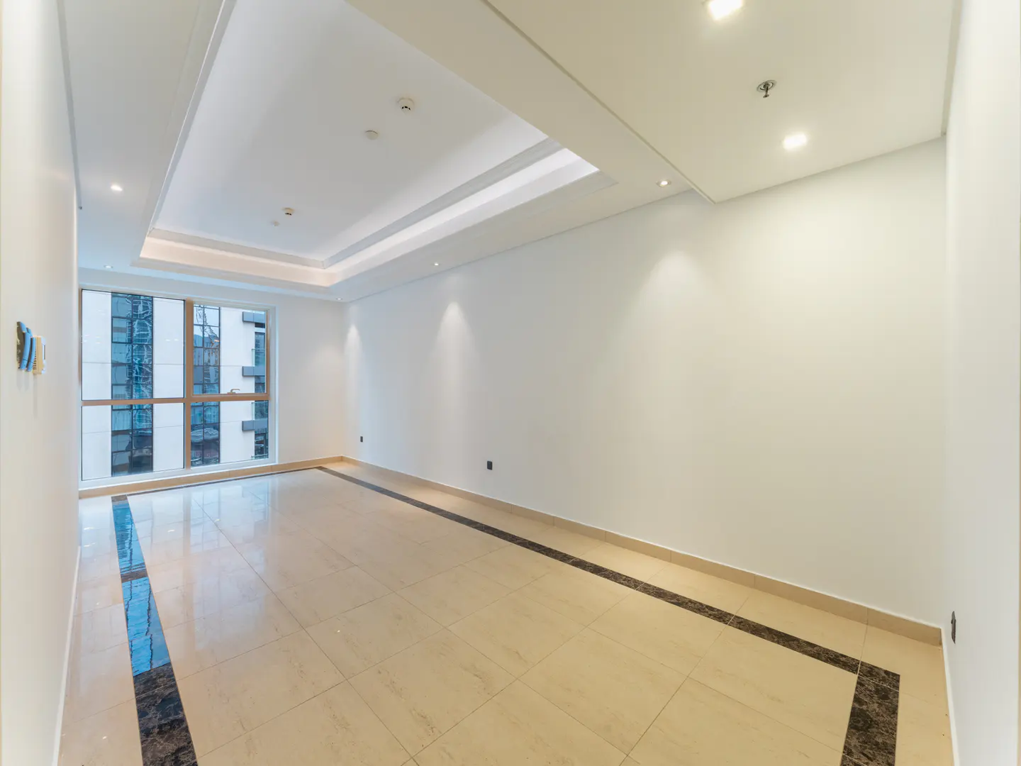 Bright, empty room with beige tile floor and white walls. A large window shows a city view. Recessed lighting in the ceiling.
