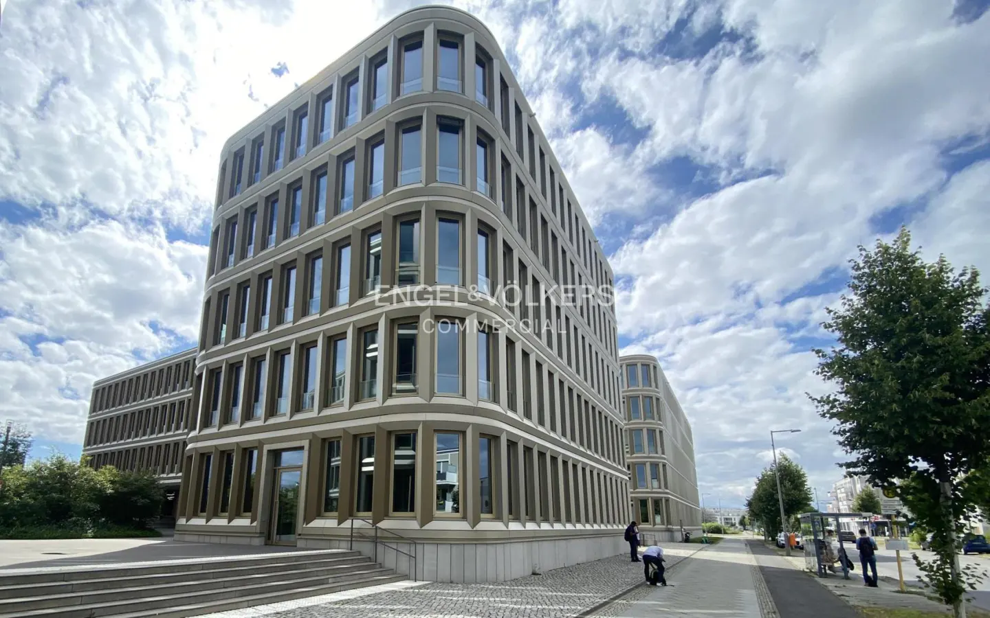 Exterior view of a modern, multi-story commercial building with many windows under a cloudy sky. The Engel & Völkers Commercial logo is visible.