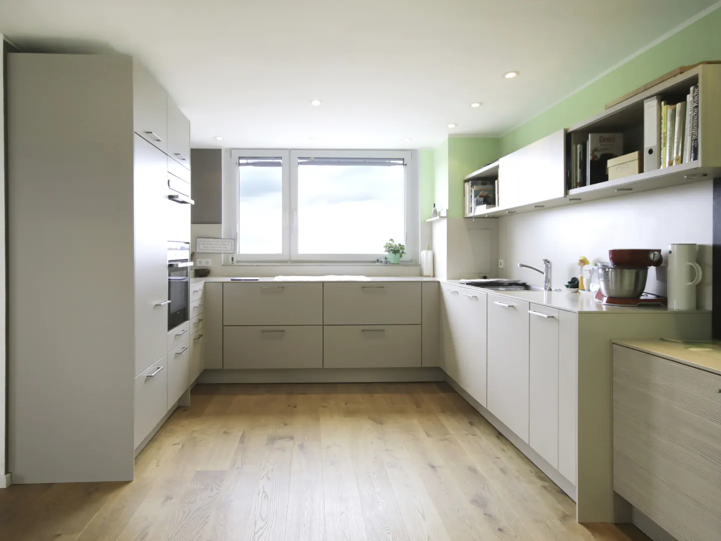 Bright kitchen with light wood floors, white cabinets, and a large window. A red stand mixer sits on the counter.