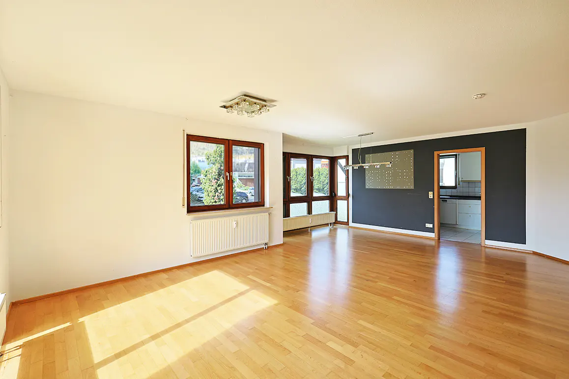 Bright, empty room with hardwood floors, white walls, and dark accent wall. Windows with brown frames let in natural light. A glimpse of the kitchen is visible.