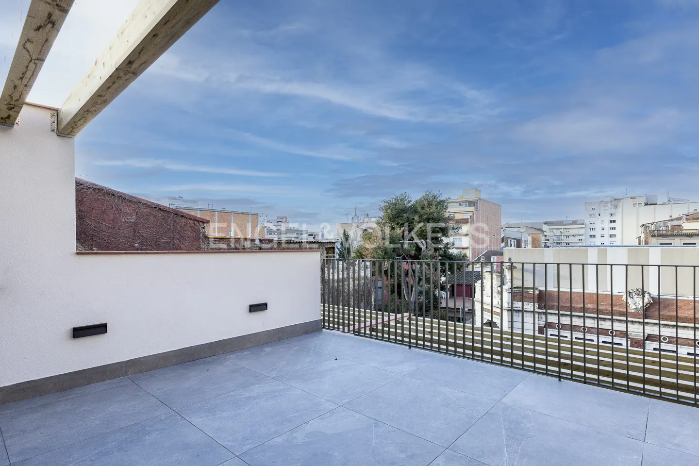 Wide shot of a rooftop terrace with gray tile, white walls, and a black metal railing overlooking a city with a cloudy blue sky.