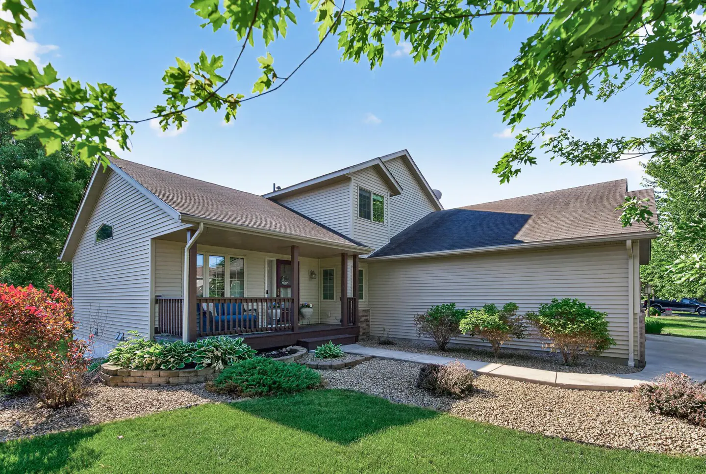 Two-story beige vinyl-sided house with a covered front porch, gravel landscaping, and green lawn under a blue sky.