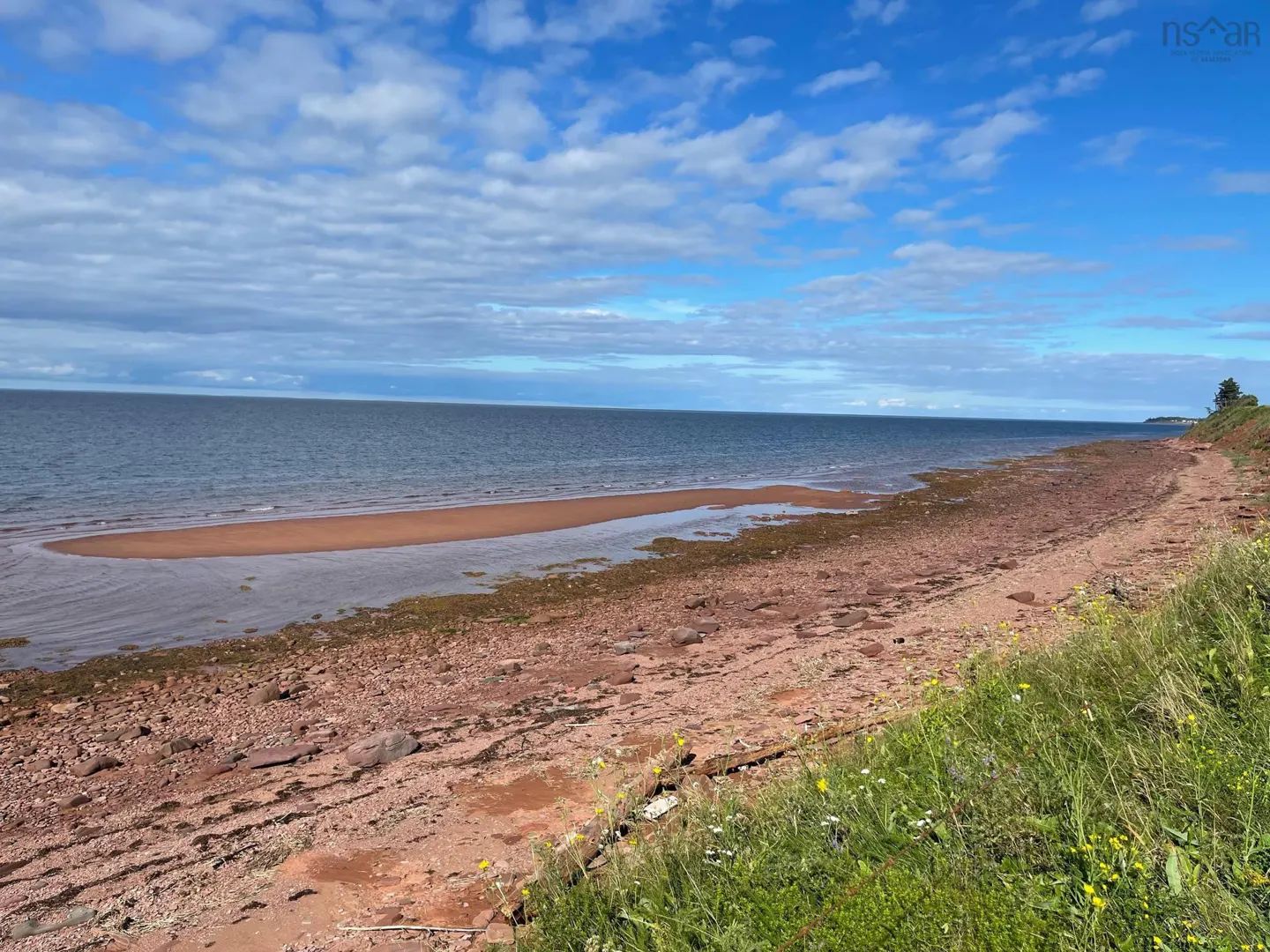 Ocean view of a red sand beach with green grass in the foreground under a blue sky with white clouds.