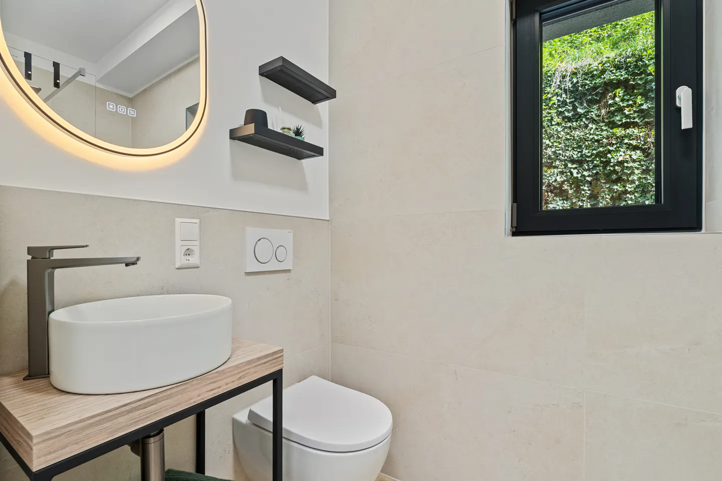 Modern bathroom with a white sink, black faucet, and a toilet. A black window shows green foliage outside.
