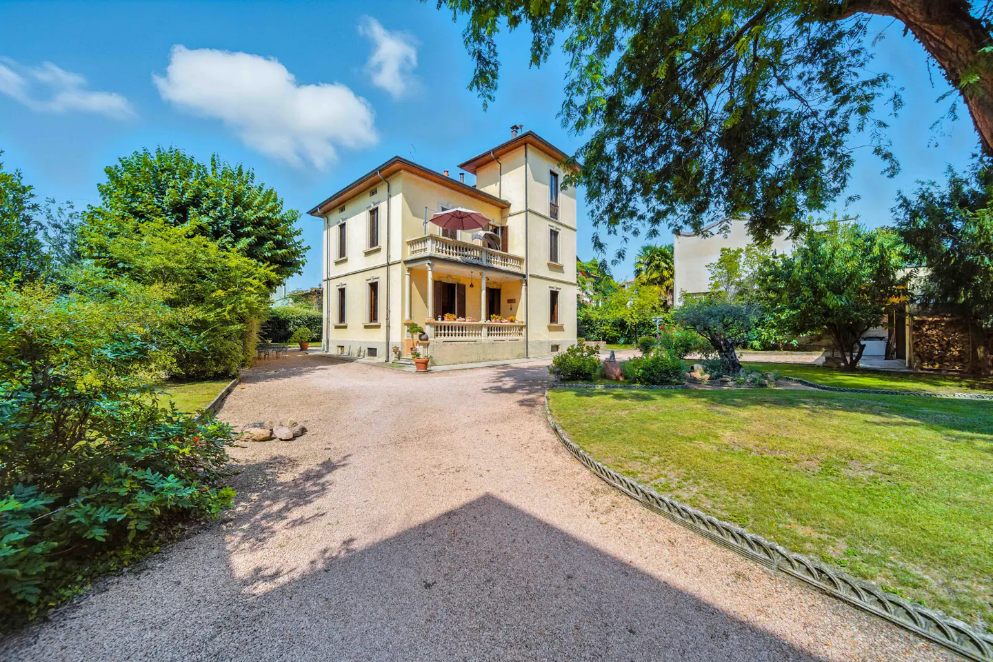 Two-story beige house with a balcony and a gravel driveway, surrounded by green trees and grass under a blue sky.