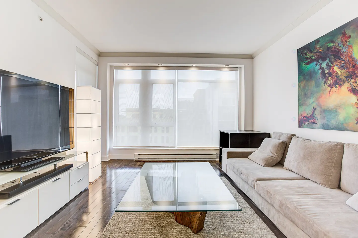 Living room with a beige sofa, glass coffee table with a wood base, and a large abstract painting on the wall.