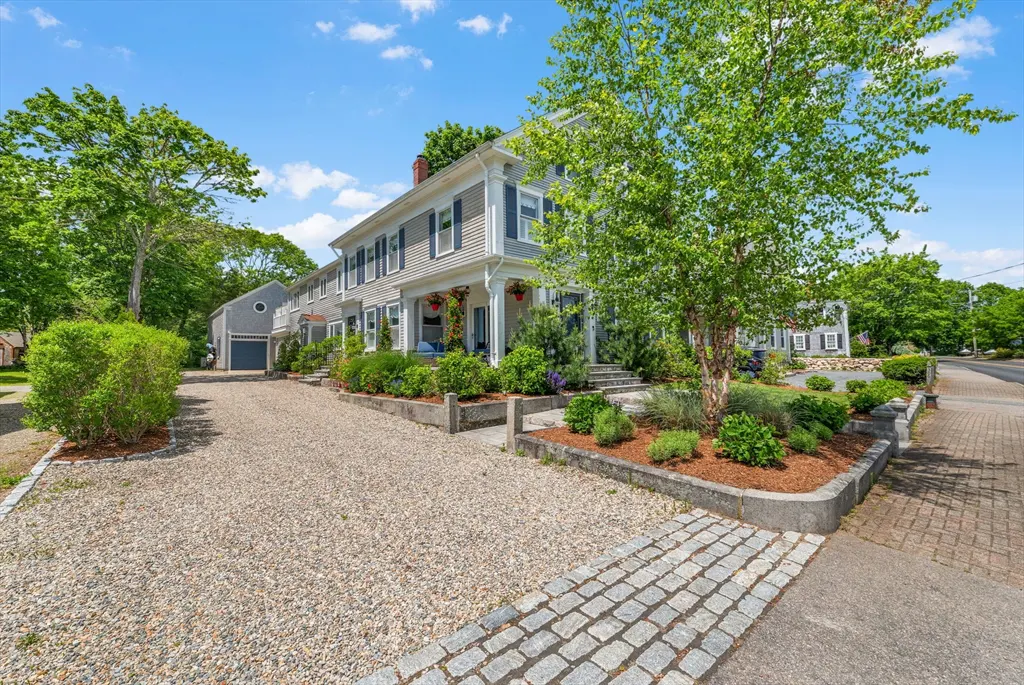A grey two-story house with blue shutters, a gravel driveway, and a landscaped front yard under a bright blue sky.