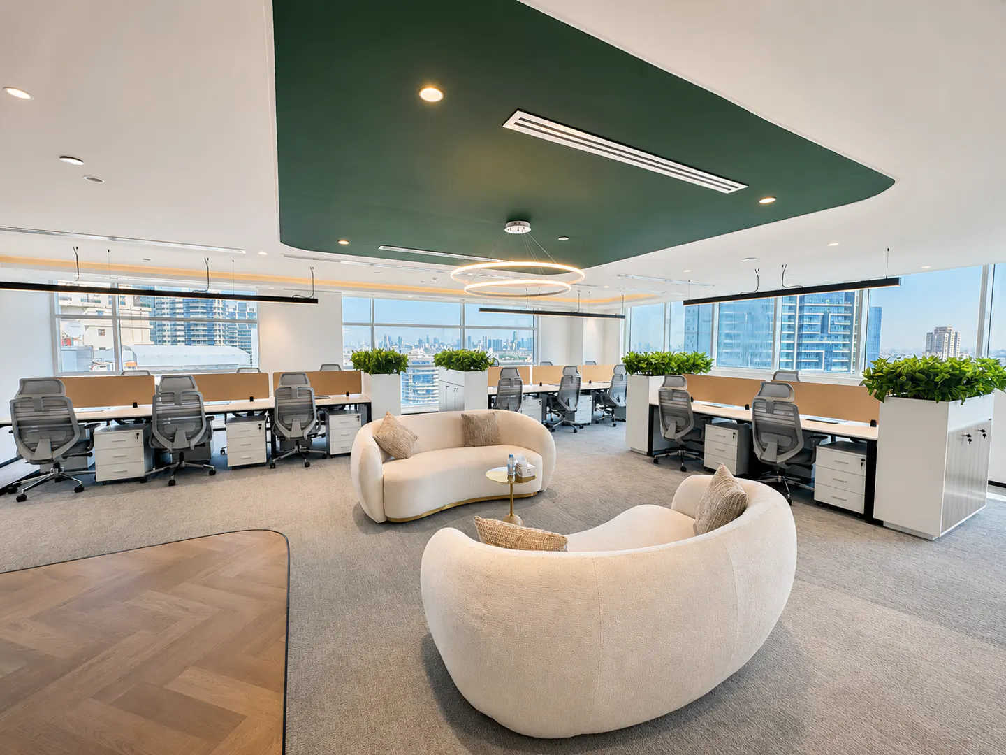 Modern office with two curved white sofas, workstations, green ceiling accents, and large windows overlooking a city skyline.