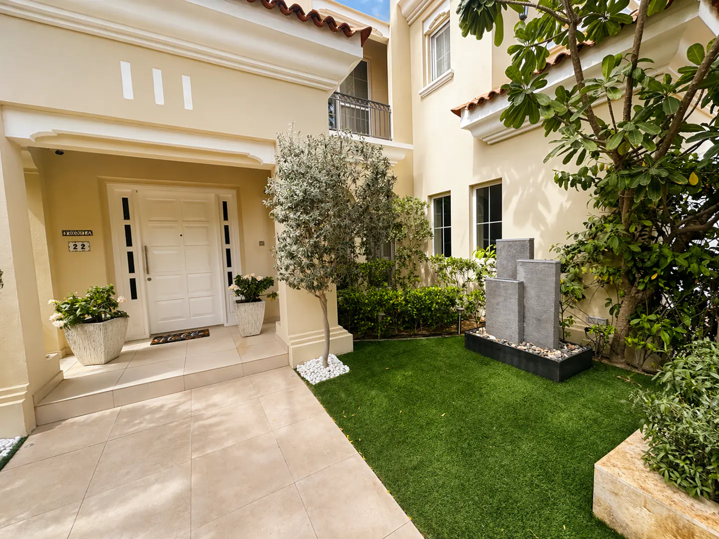 Beige two-story house with a white front door, potted plants, a small tree, and a modern stone fountain on green grass.