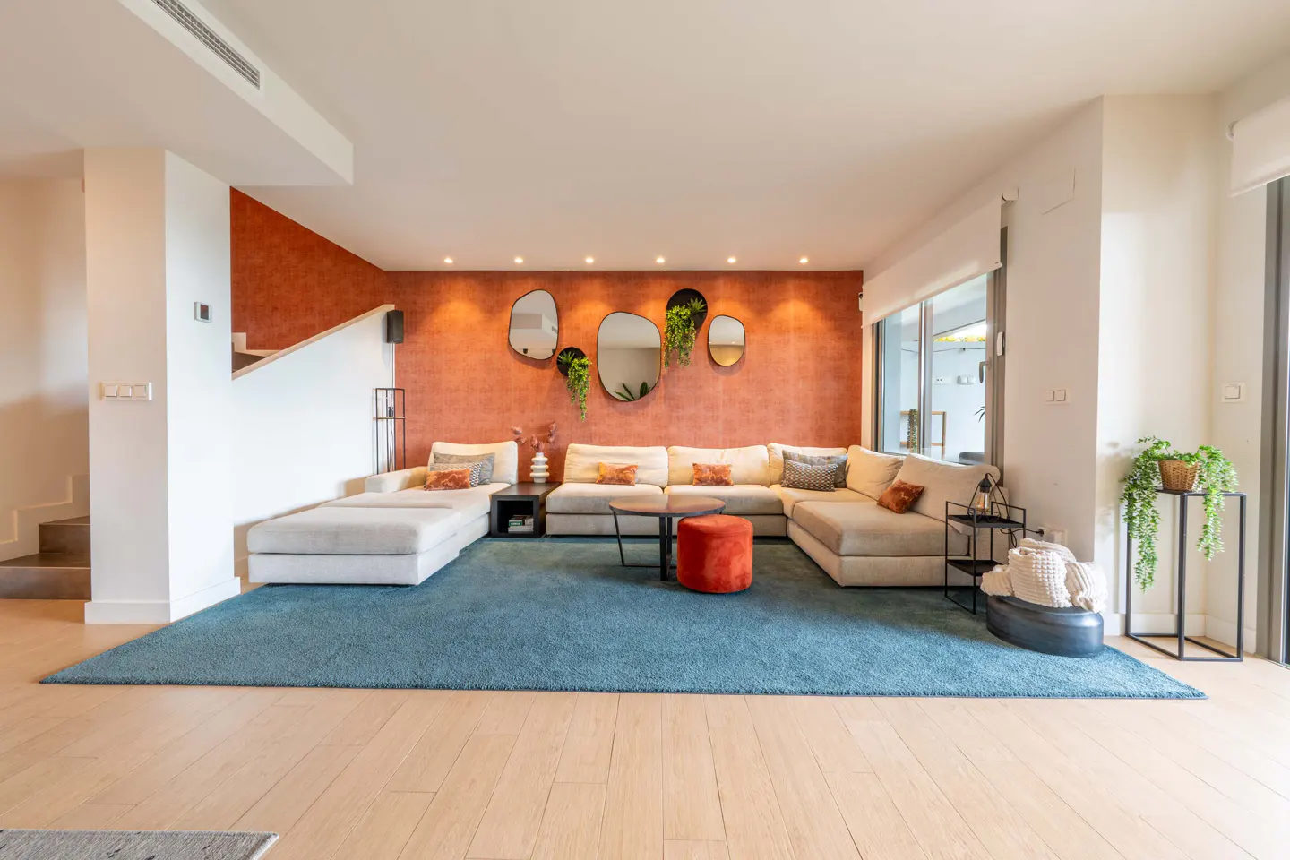 Bright living room with a large sectional sofa, blue rug, and orange accent wall with decorative mirrors and plants.