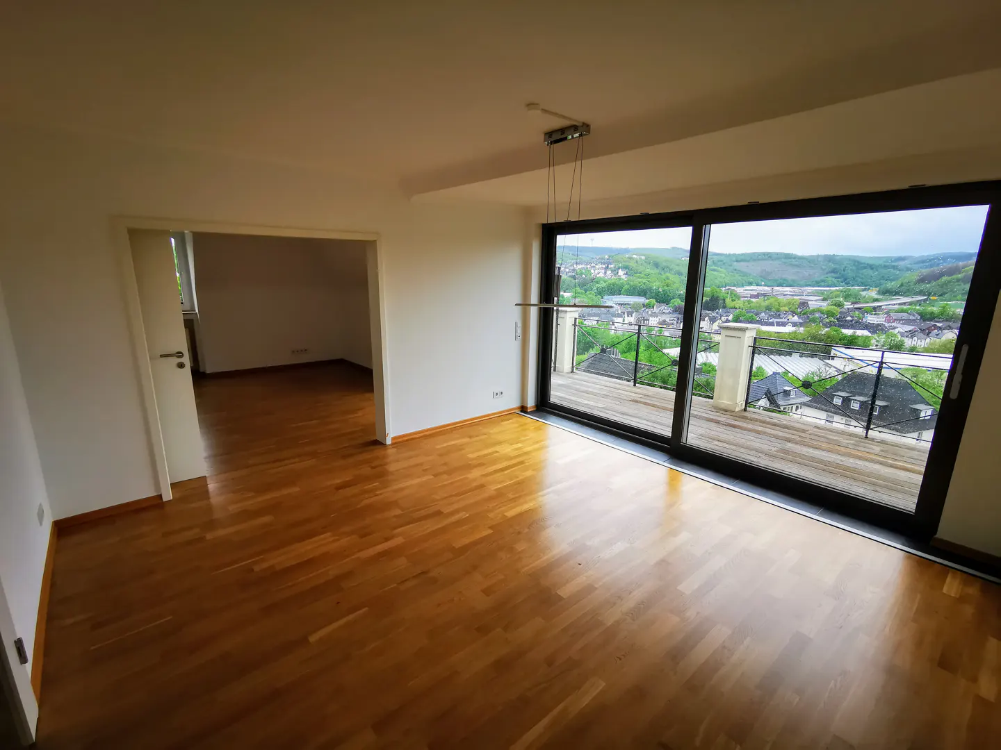An empty room with wood floors and white walls. A large sliding glass door leads to a balcony with a view of a green valley.