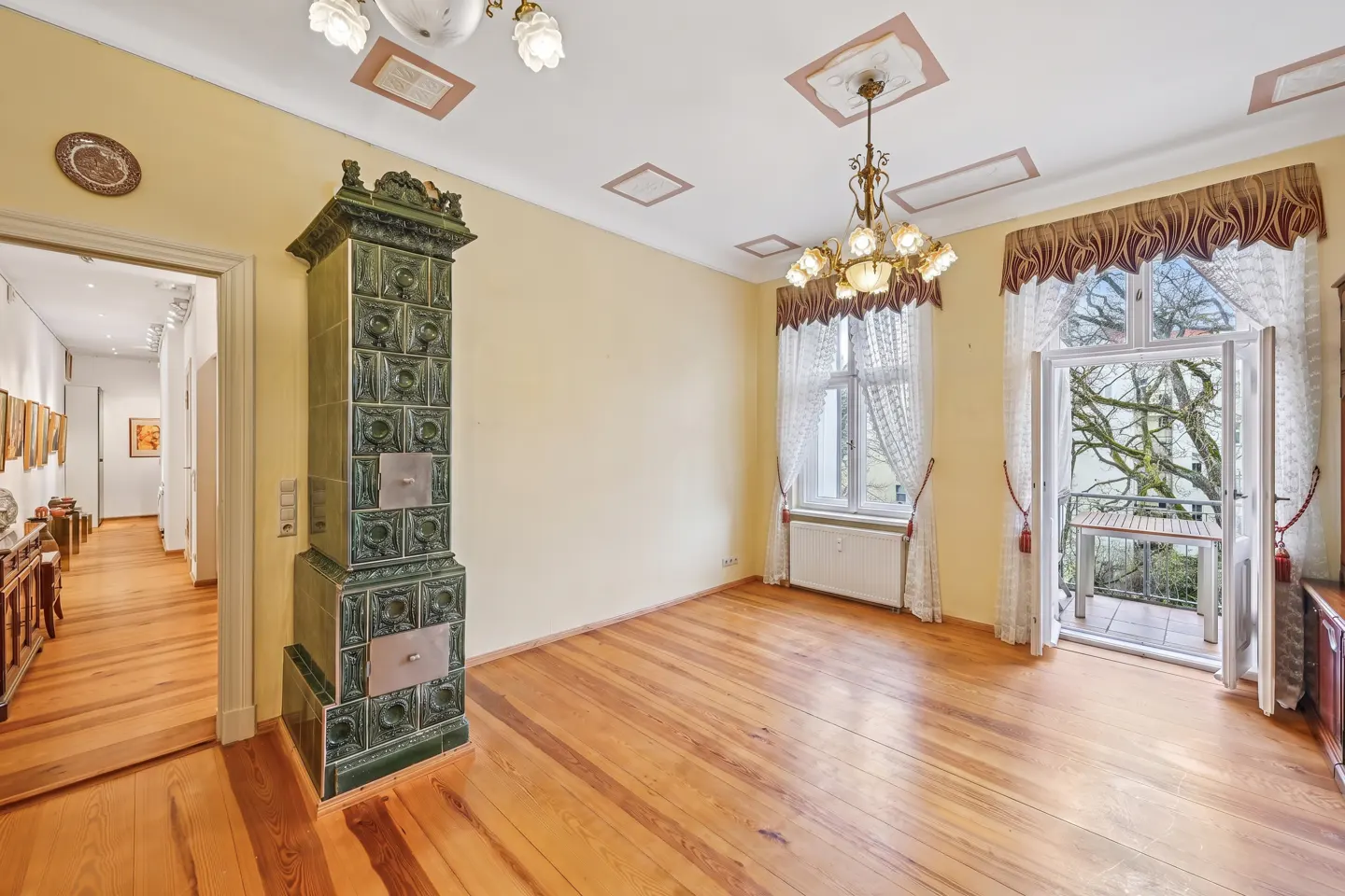 A room with yellow walls, wood floors, a green tiled stove, and a balcony with open doors.