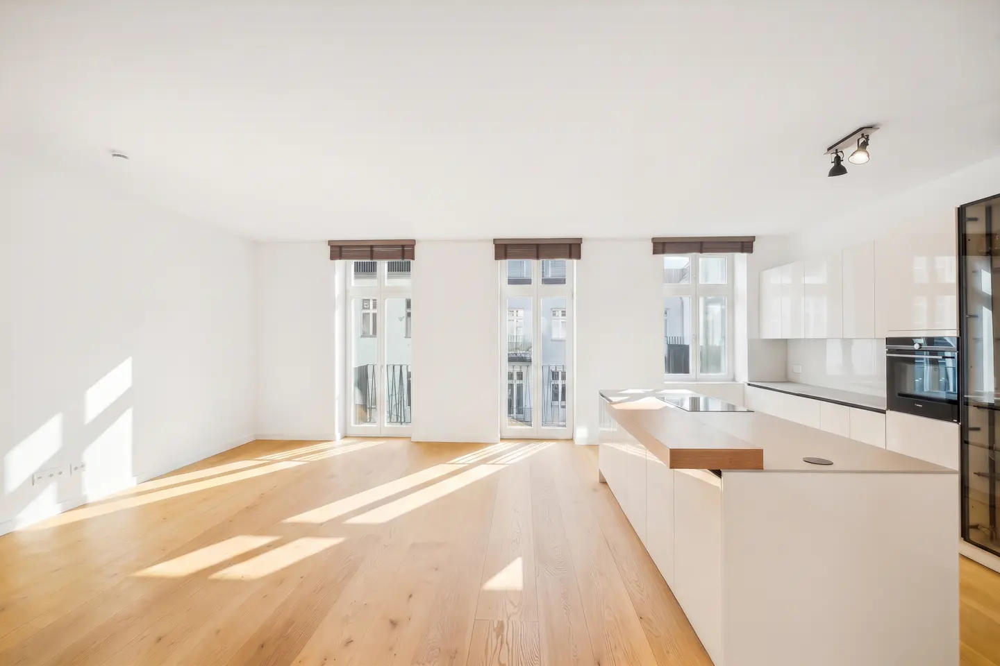 Bright, modern apartment with hardwood floors, white walls, and a kitchen island. Three windows with brown shades let in sunlight.