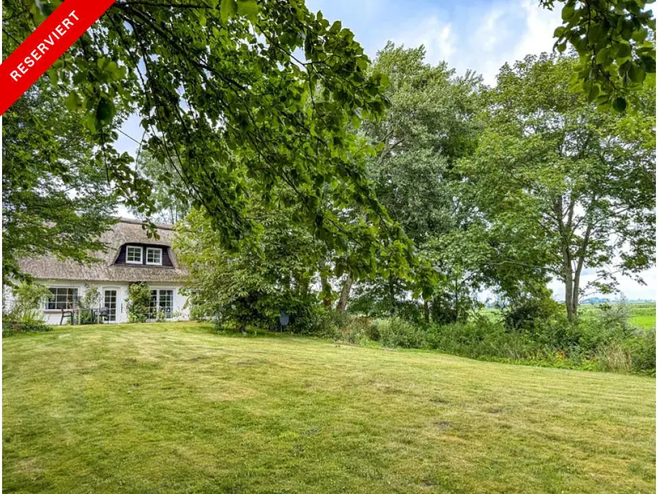 White cottage with a thatched roof, surrounded by green trees and a large lawn. "Reserviert" banner in the upper left corner.