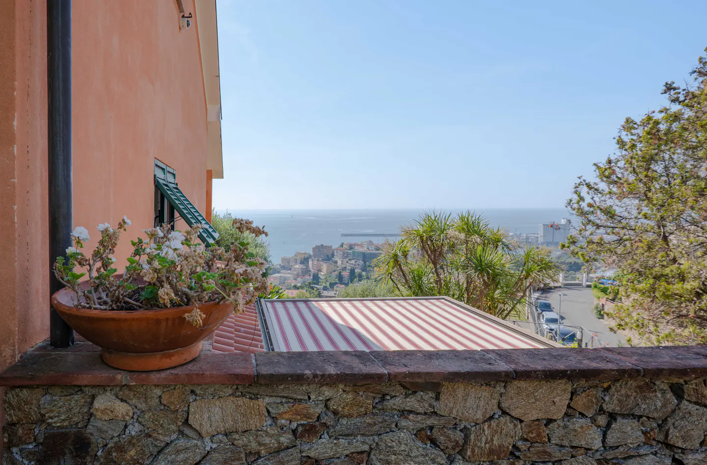 Balcony view of the sea and town. A terracotta pot with white flowers sits on a stone wall. An orange building is to the left.