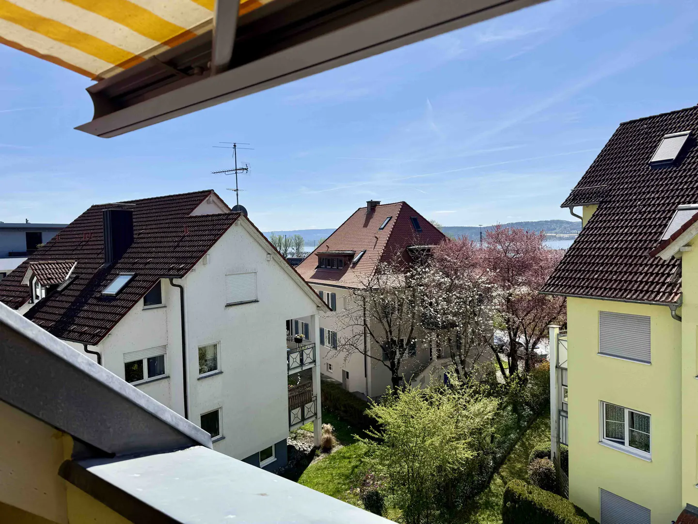 View from a balcony with a yellow and white striped awning, overlooking houses, trees, and a lake under a blue sky.