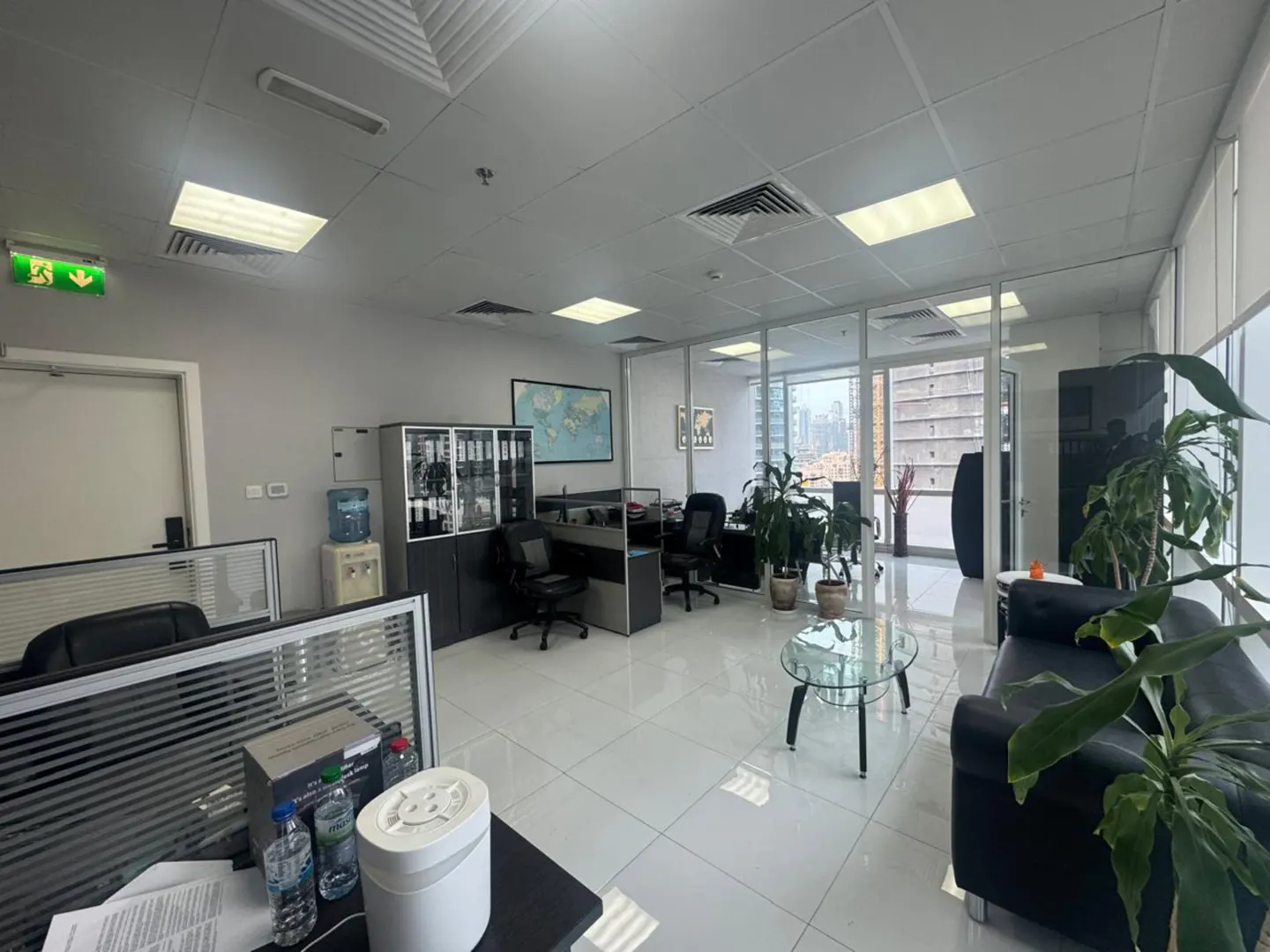 Bright office space with desks, chairs, cabinets, and a black sofa. Large windows offer a city view. Plants add a touch of nature.