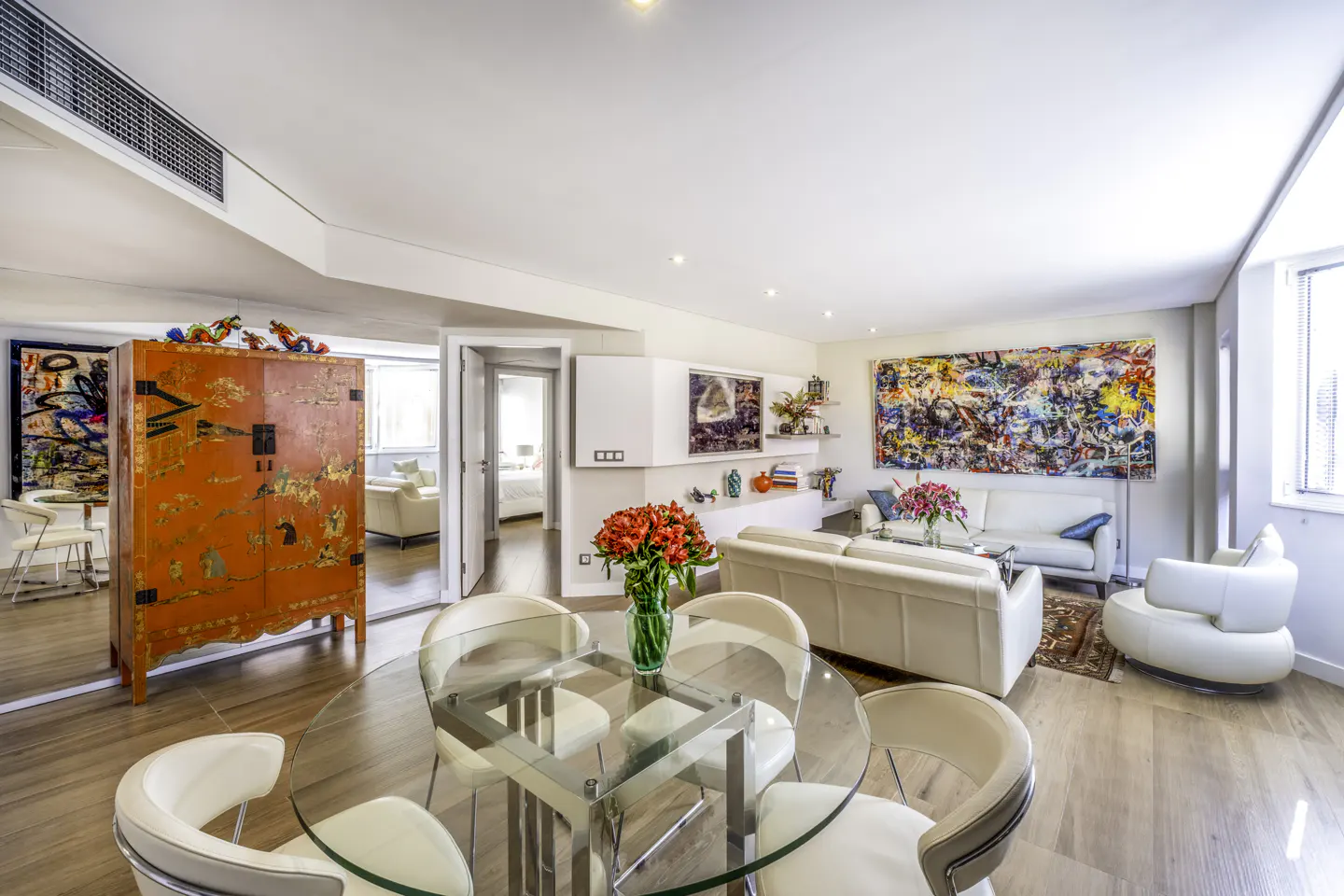 Bright, open-concept living space with a glass table, white leather furniture, and colorful art. A red cabinet adds a pop of color.