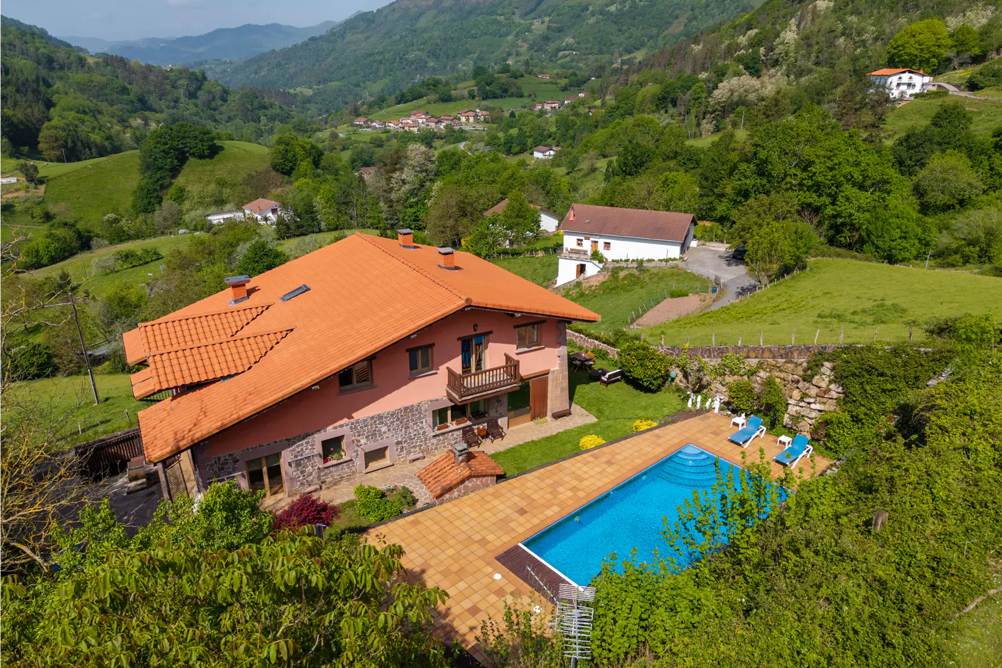 Aerial view of a pink house with an orange roof and a blue pool, surrounded by green trees and mountains.