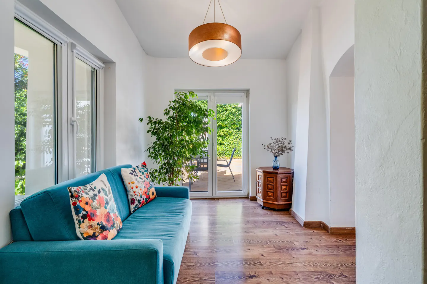 Bright living room with a teal sofa, floral pillows, wood floors, and a view to a green backyard through glass doors.