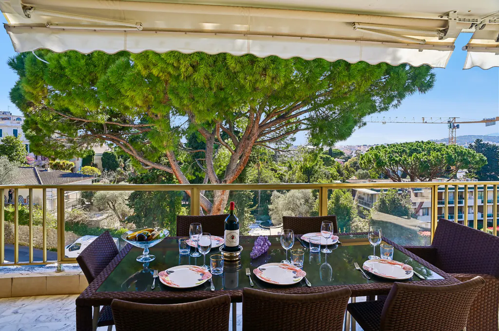 Outdoor dining area with a table set for a meal, featuring wine, plates, and a scenic view of trees and buildings.
