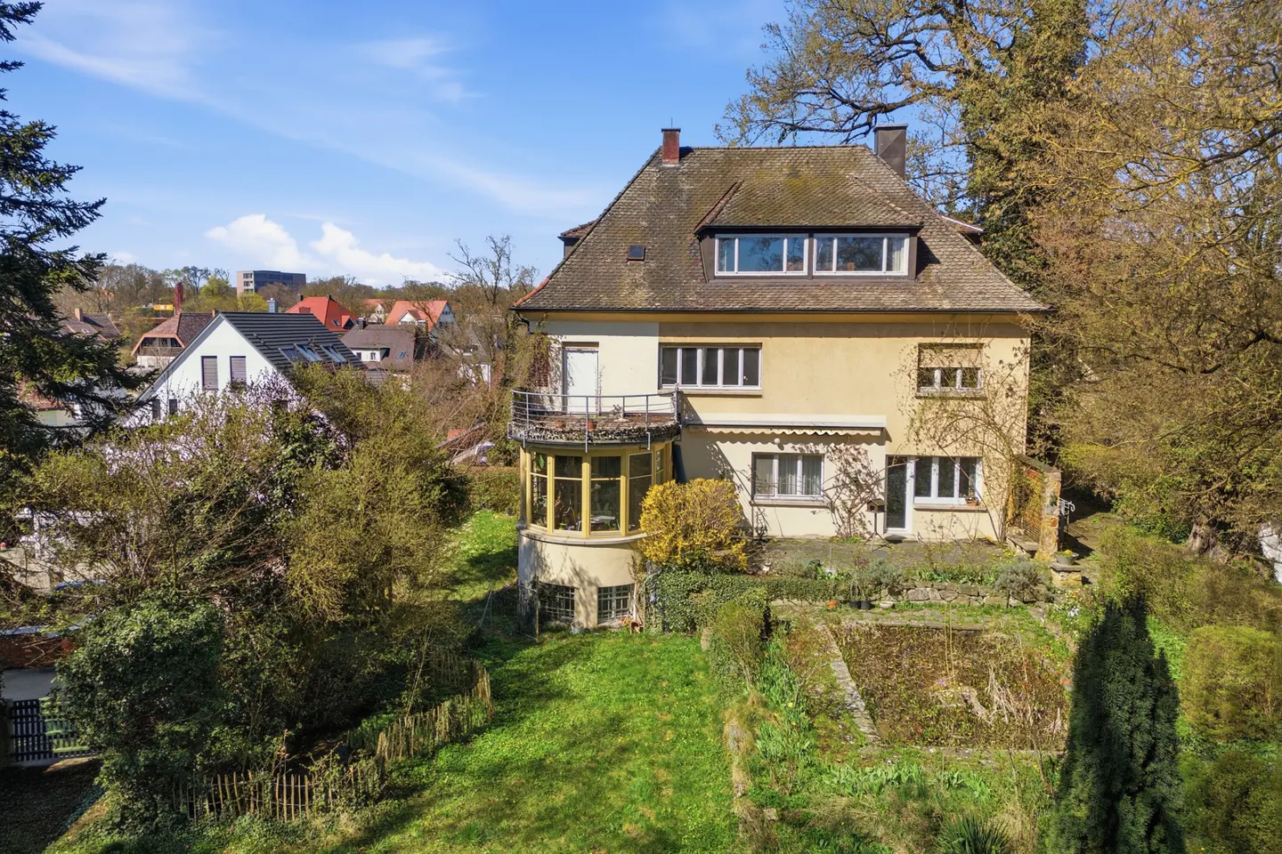 Two-story yellow house with a brown roof, balcony, and sunroom surrounded by trees and a green lawn.