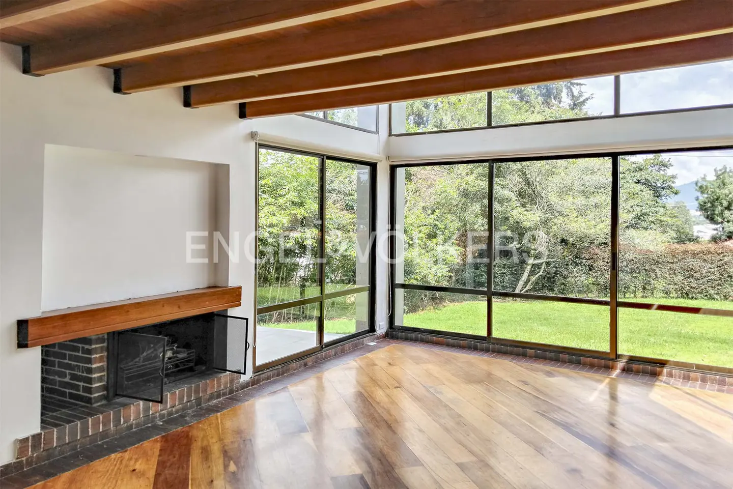 Bright living room with wood floors, exposed beams, and a brick fireplace. Large windows offer views of a green lawn and trees.