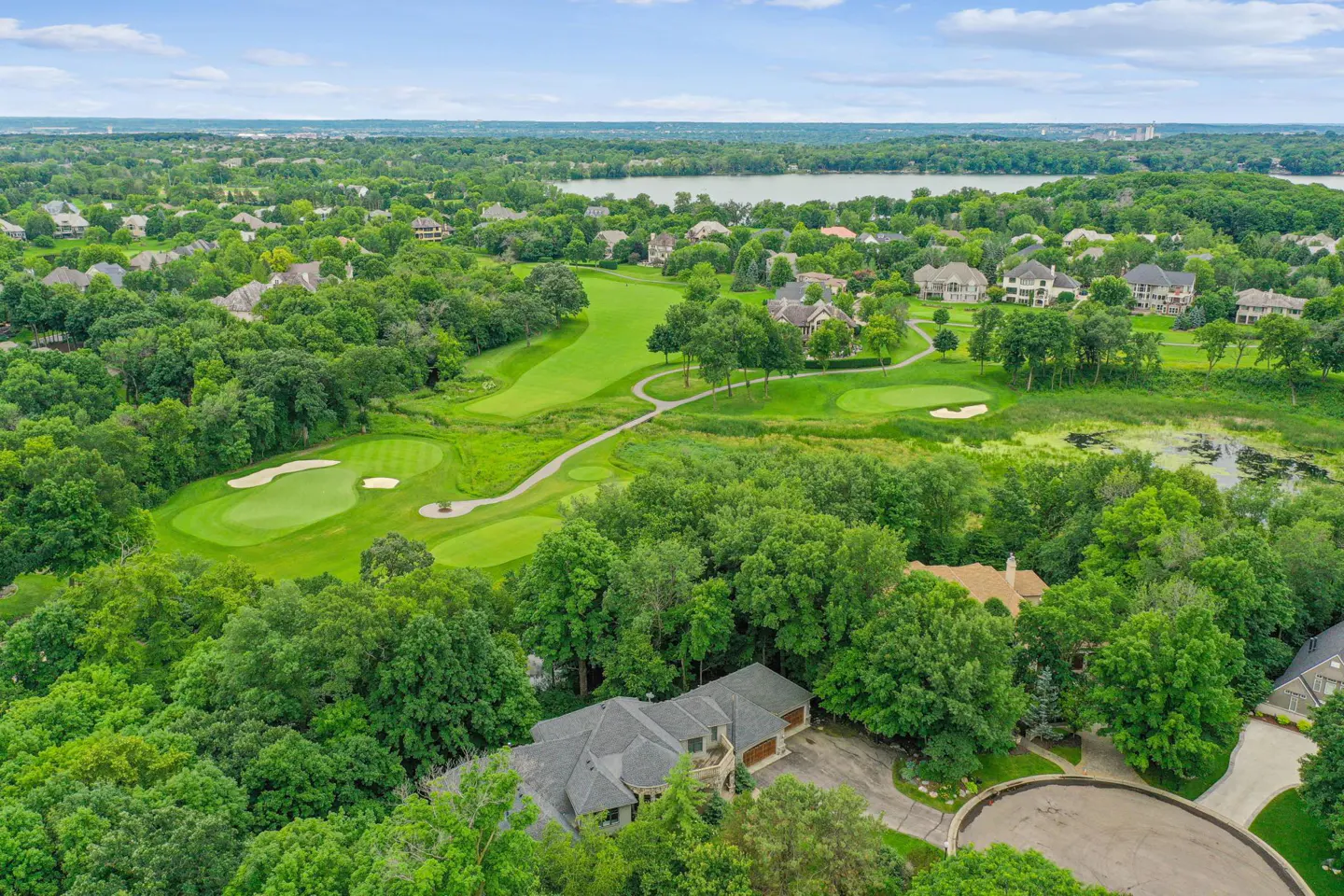 Aerial view of a luxury home nestled in lush trees overlooking a sprawling green golf course and a distant lake.