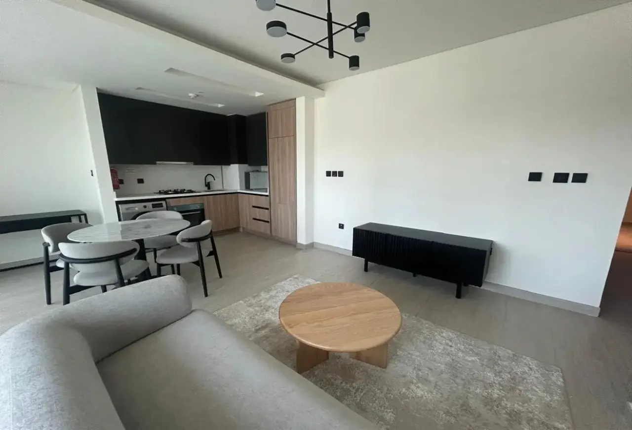 Modern open-plan living area with a grey sofa, wooden coffee table, dining set, and a sleek black and wood-toned kitchen.