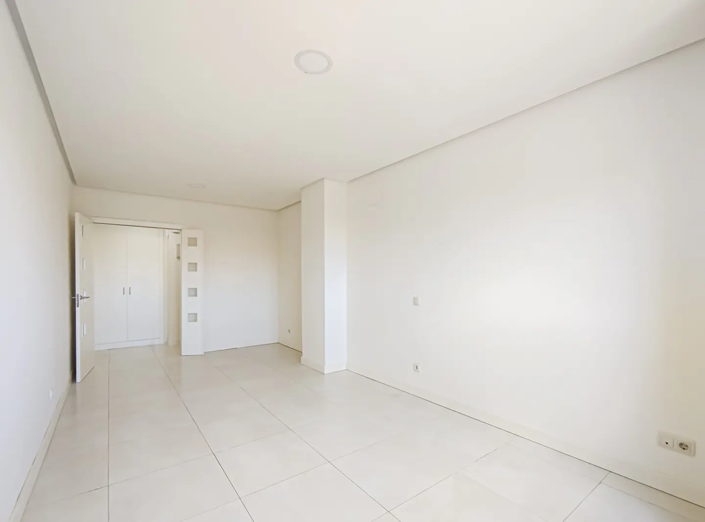 Bright, empty room with white walls and tile floor. A closet with double doors is visible in the background.