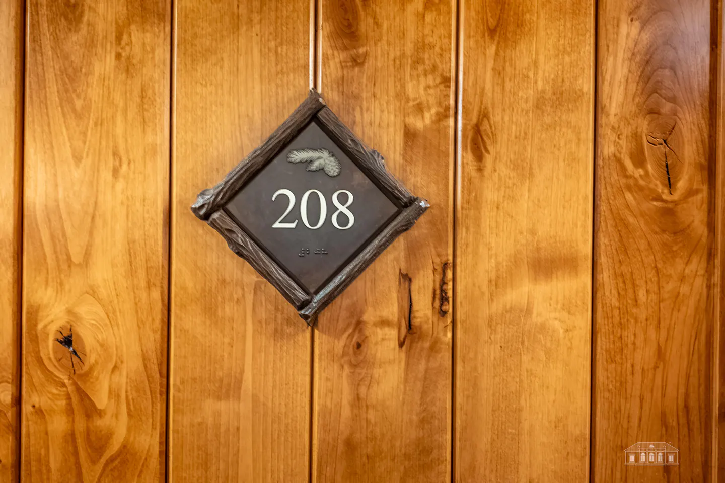 Wooden door with a diamond-shaped sign reading "208" in white numbers, with braille below and a feather at the top.