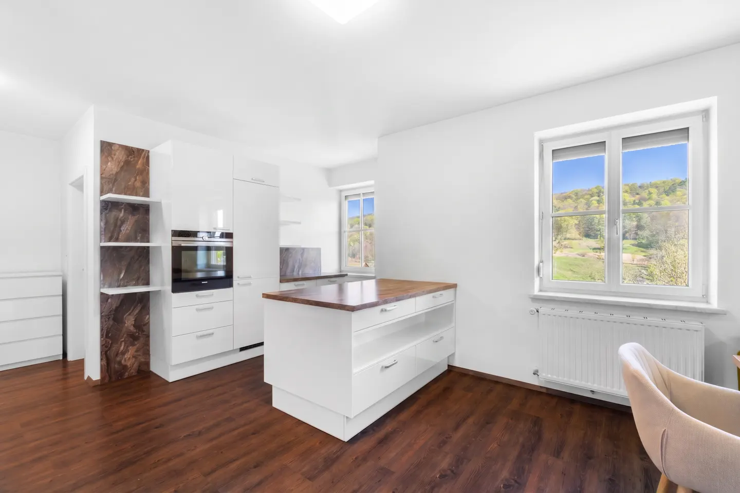 Bright, modern kitchen with white cabinets, wood floors, and a central island. A window shows a green, hilly landscape.