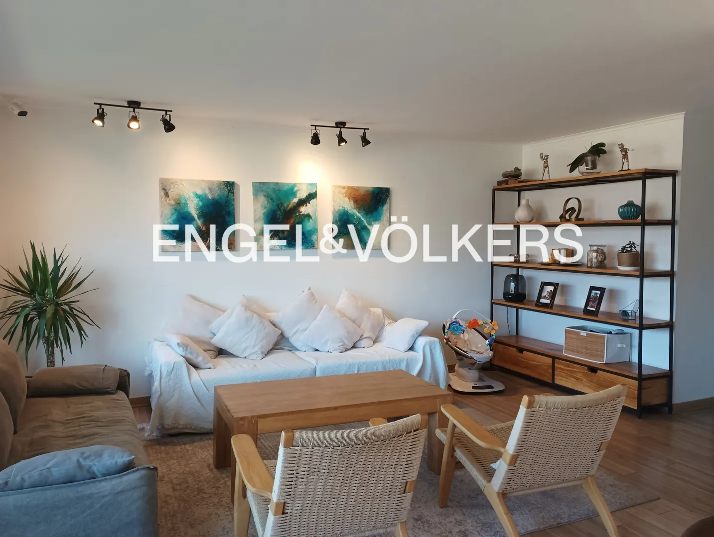 Living room with white walls, light wood floors, and modern furniture. A white sofa with pillows sits under abstract art. A wooden coffee table and woven chairs are in front. A bookshelf is on the right.