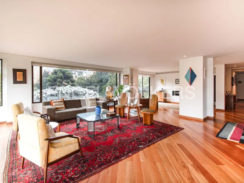 Bright living room with wood floors, a red rug, and a large window overlooking trees. A gray sofa and yellow armchairs provide seating.
