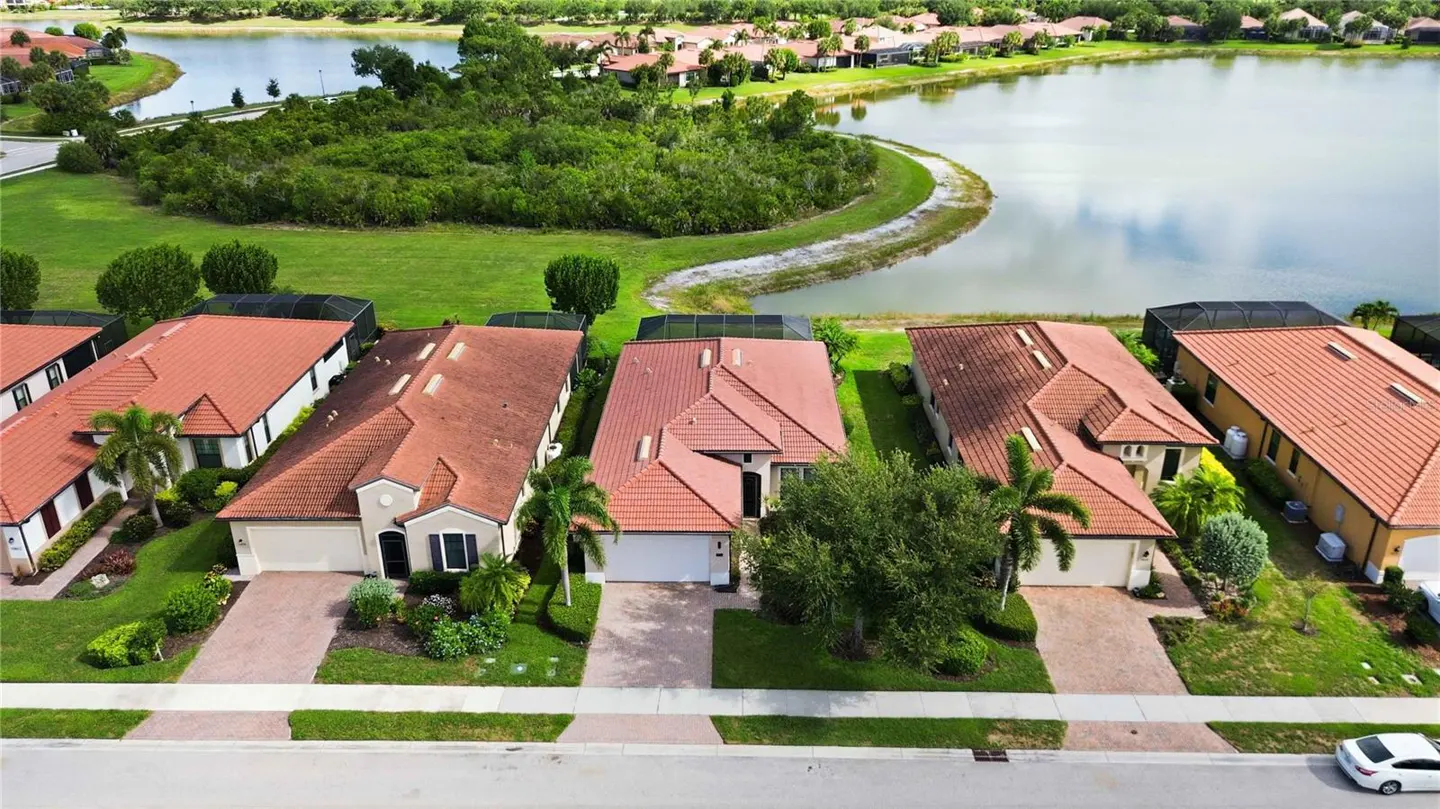 Aerial view of suburban homes with red tile roofs, paved driveways, and lush landscaping overlooking a scenic lake.