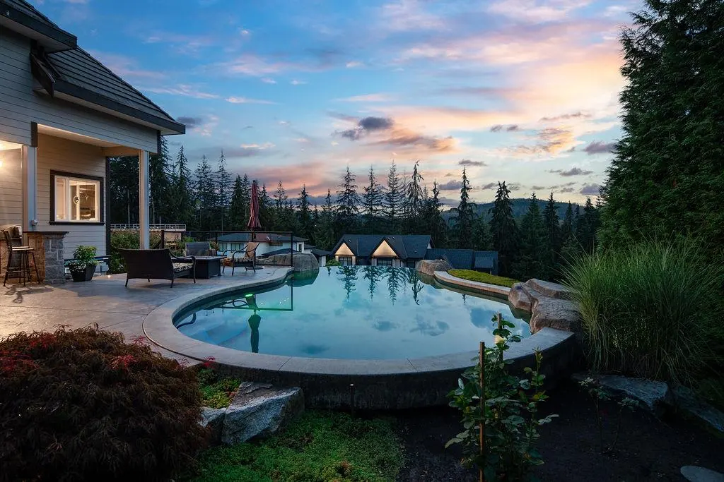 Outdoor pool reflects a colorful sunset sky. Patio with seating, trees, and houses in the background.