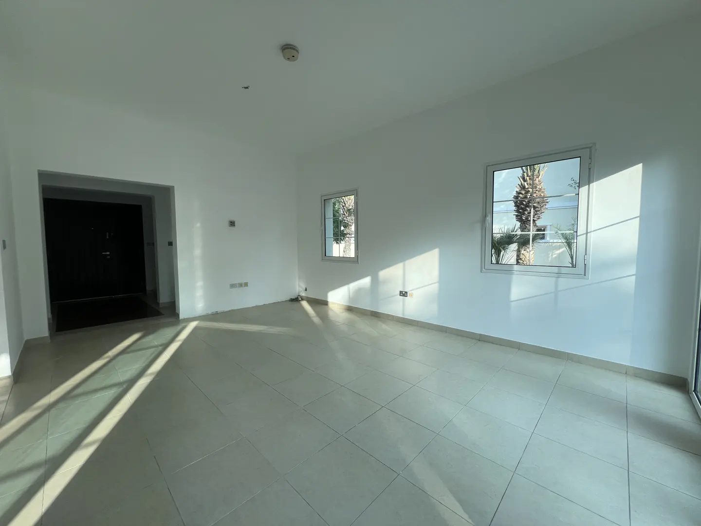 Bright, empty room with white walls, tile floor, and two windows showing a palm tree outside. A dark doorway is on the left.