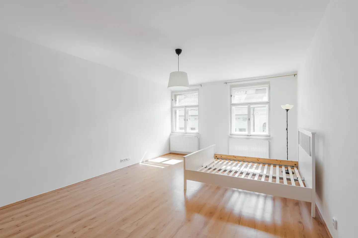 Bright, empty bedroom with white walls, wood floor, and a white bed frame. Two windows let in sunlight. A white lamp hangs from the ceiling.