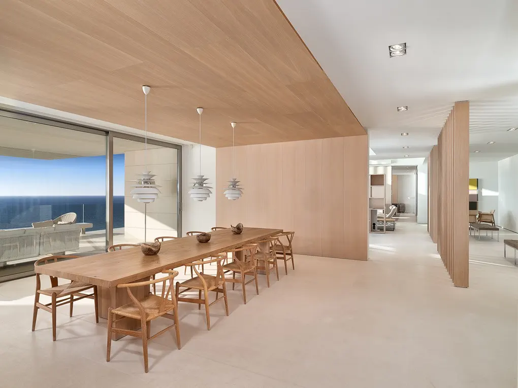 A bright, modern dining room with a long wooden table, chairs, and ocean view. Three pendant lights hang above the table.