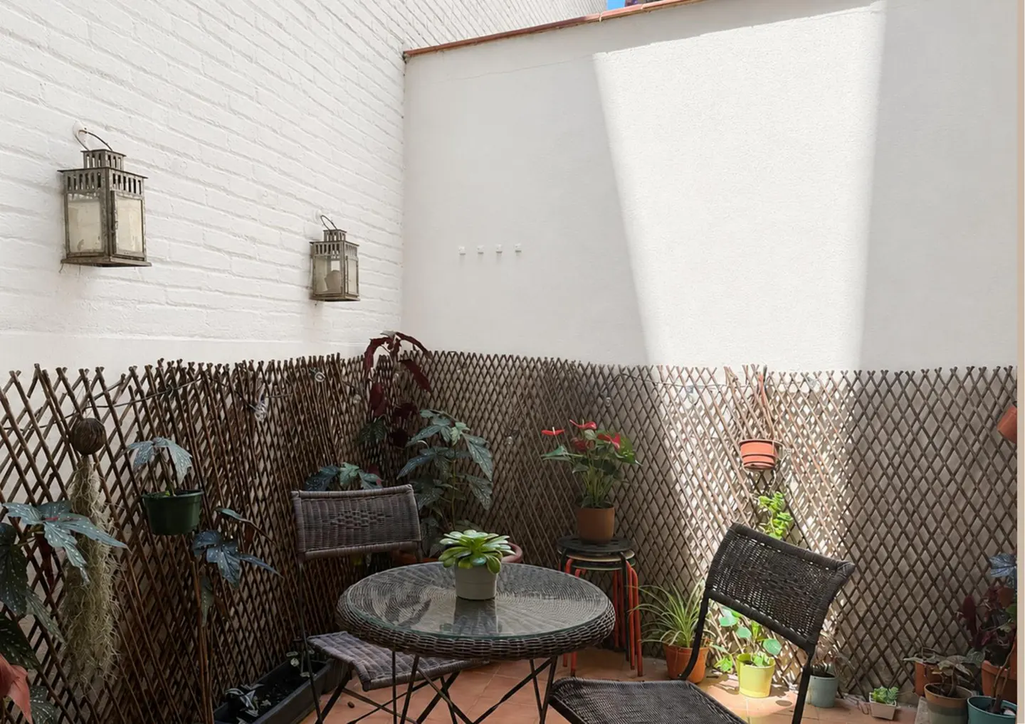 Outdoor patio with wicker furniture, plants, and lanterns on a white brick wall. A lattice fence provides privacy.