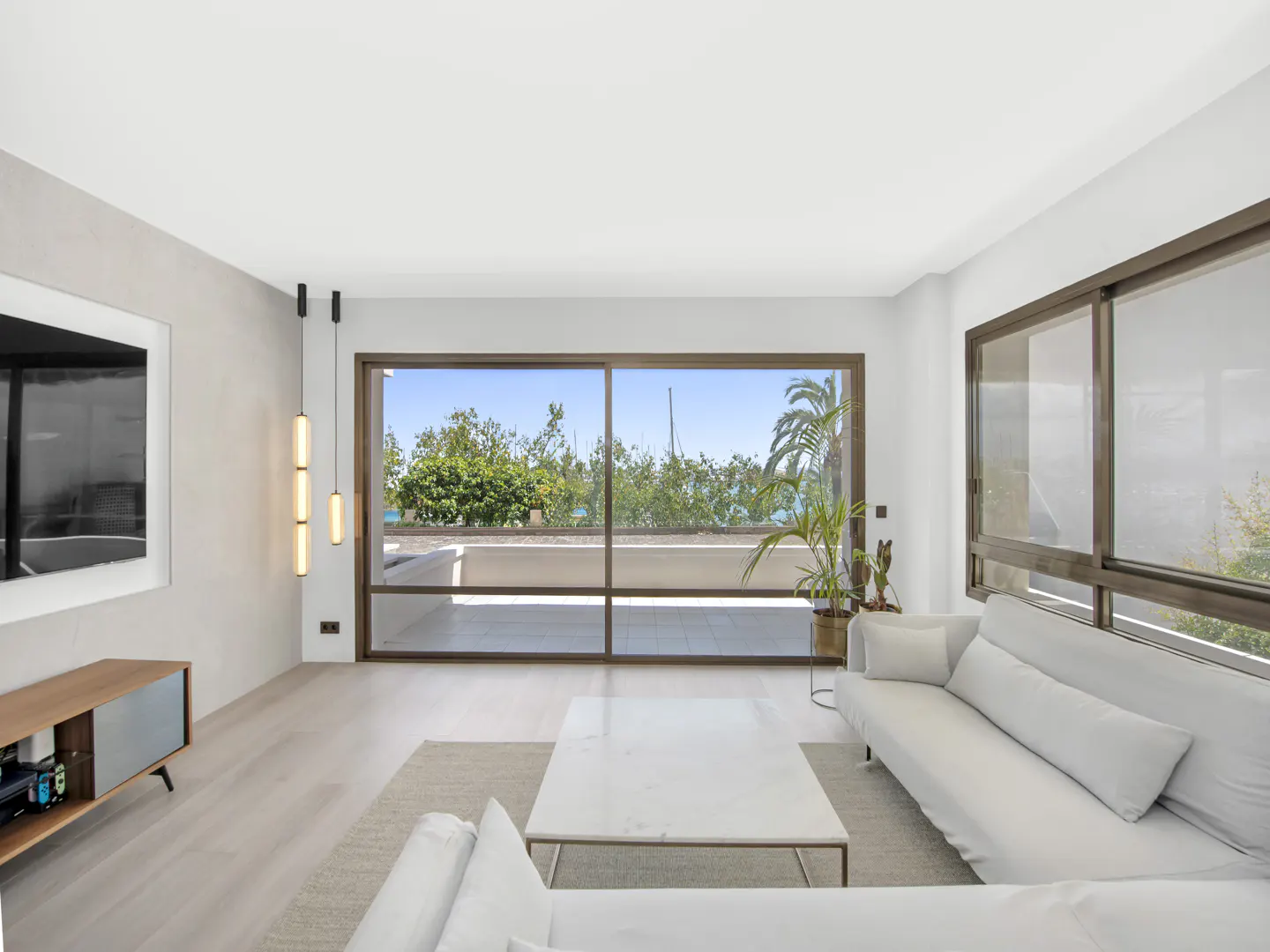 Bright living room with white sofas, a marble coffee table, and a large window overlooking a balcony with trees and ocean views.