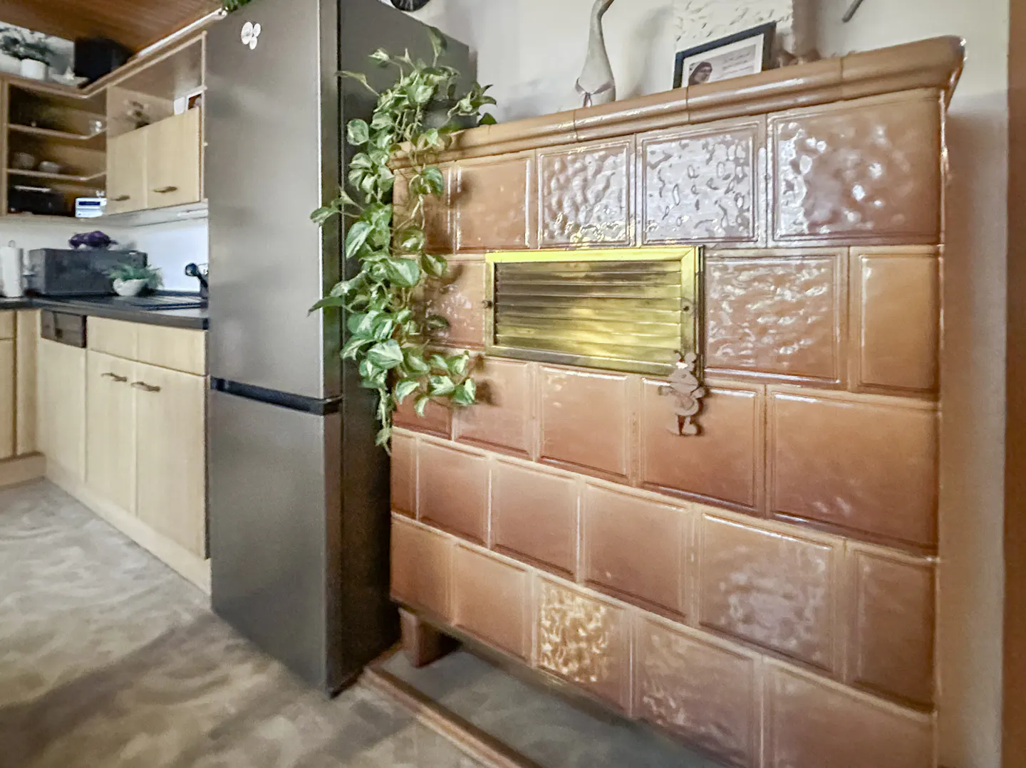 A kitchen features a large, brown tiled oven with a gold vent, next to a stainless steel refrigerator. A green plant drapes over the oven.