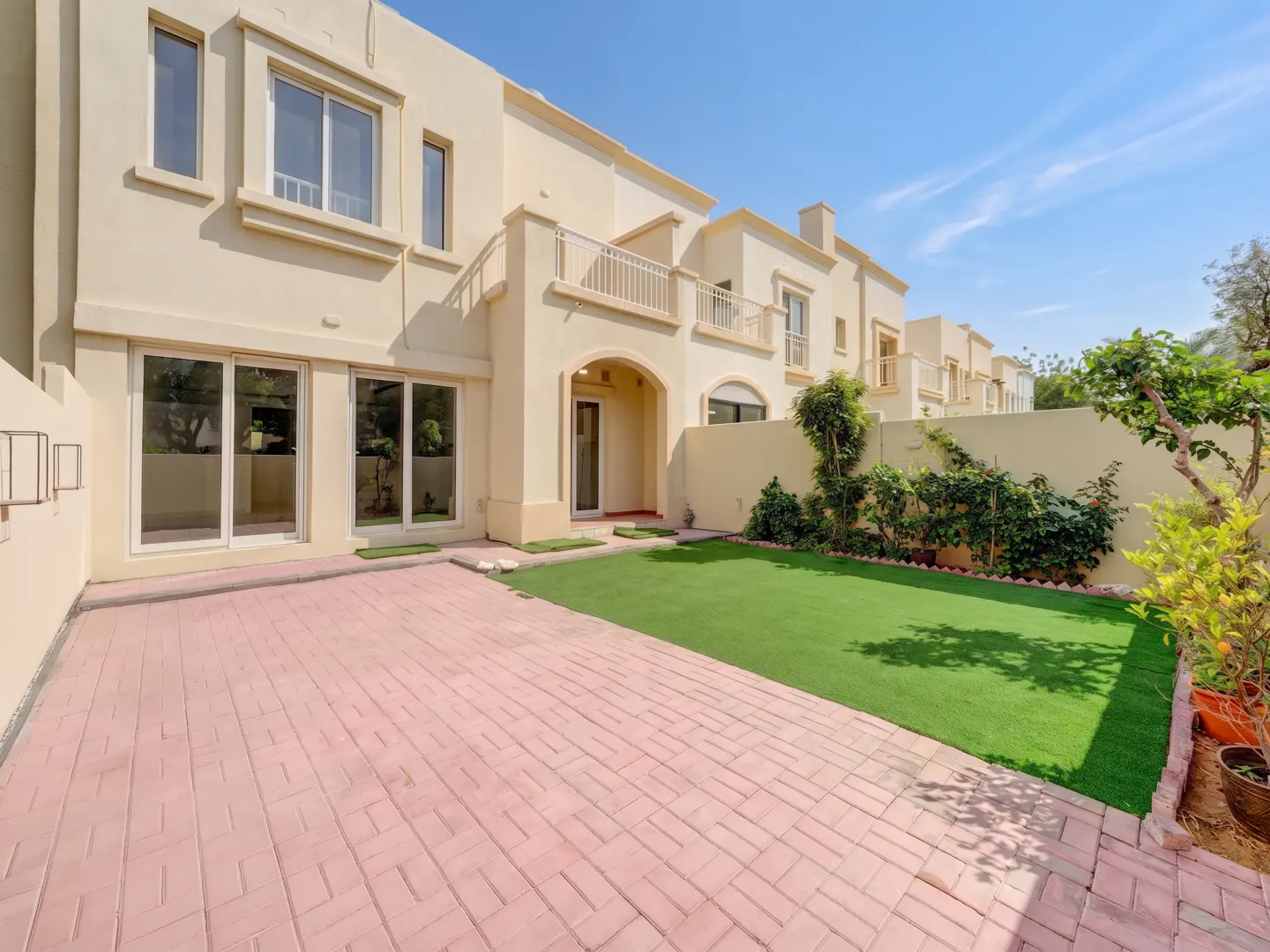 Backyard of a two-story beige house with a brick patio, green lawn, and garden with plants and trees under a blue sky.