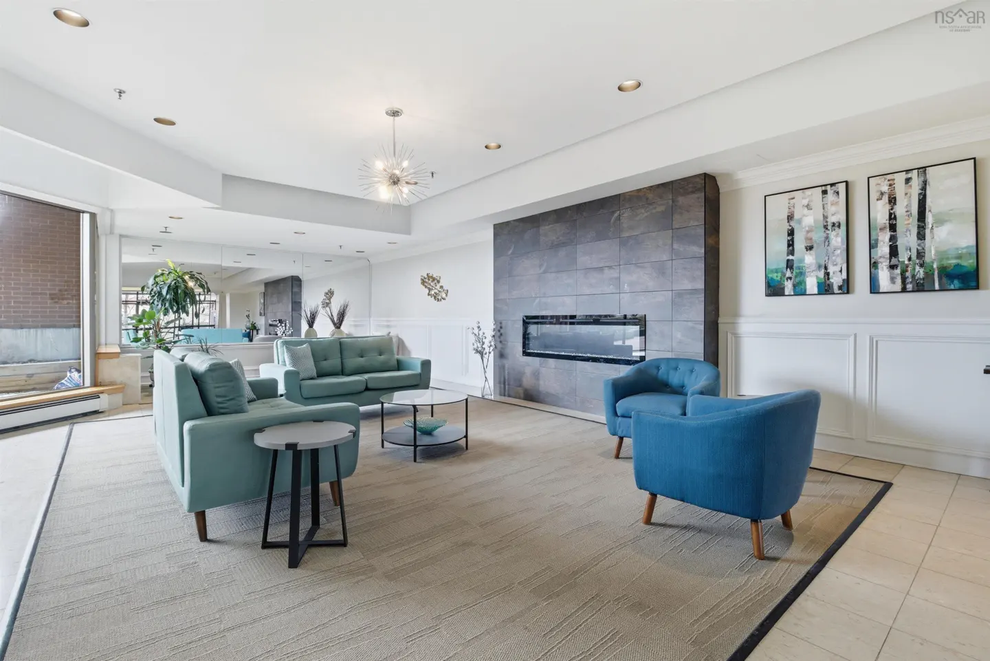 Bright, modern living room with blue sofas, chairs, and a fireplace. Artwork hangs on the white walls. A large window lets in natural light.