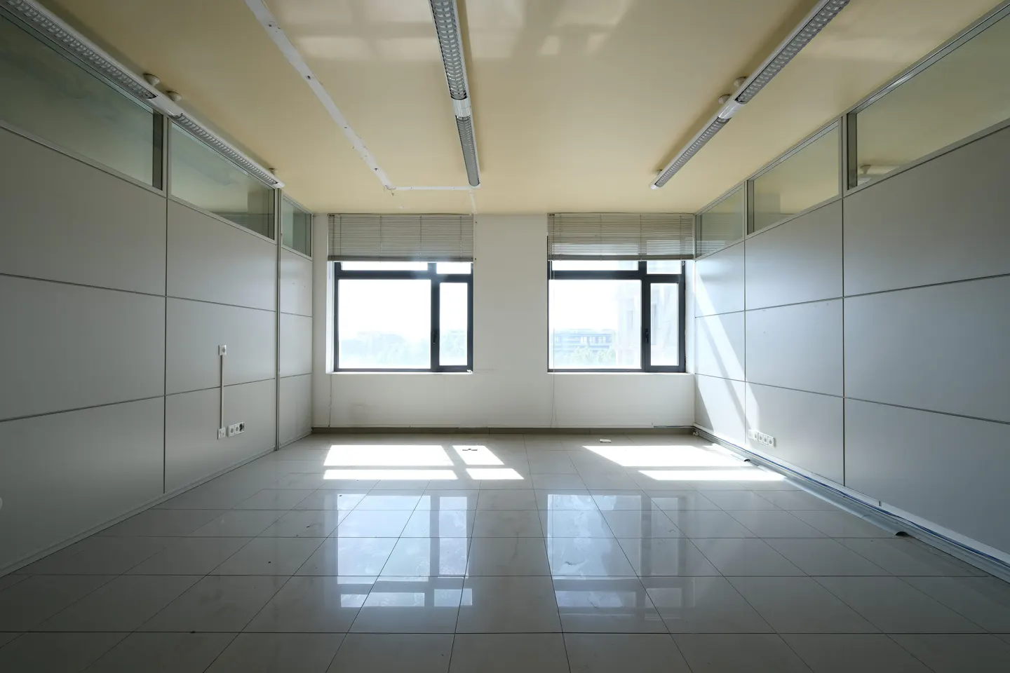 Empty office space with white walls, tile floor, and two windows with sunlight streaming in.