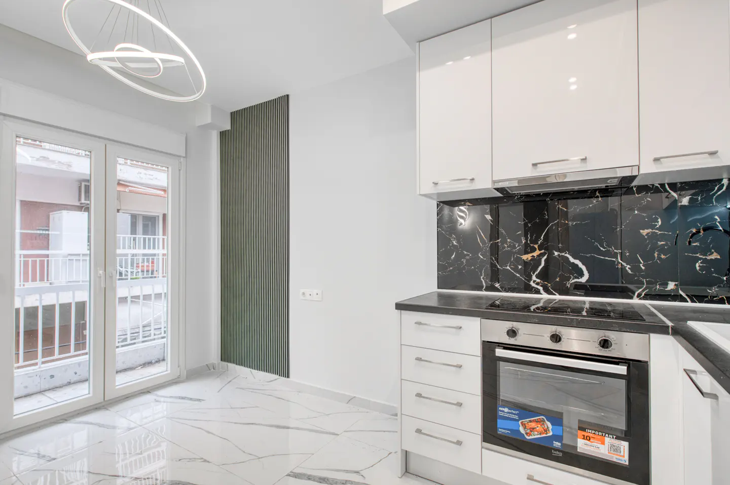 Bright kitchen with white cabinets, black marble backsplash, and white marble floor. A modern light fixture hangs above a glass door to a balcony.