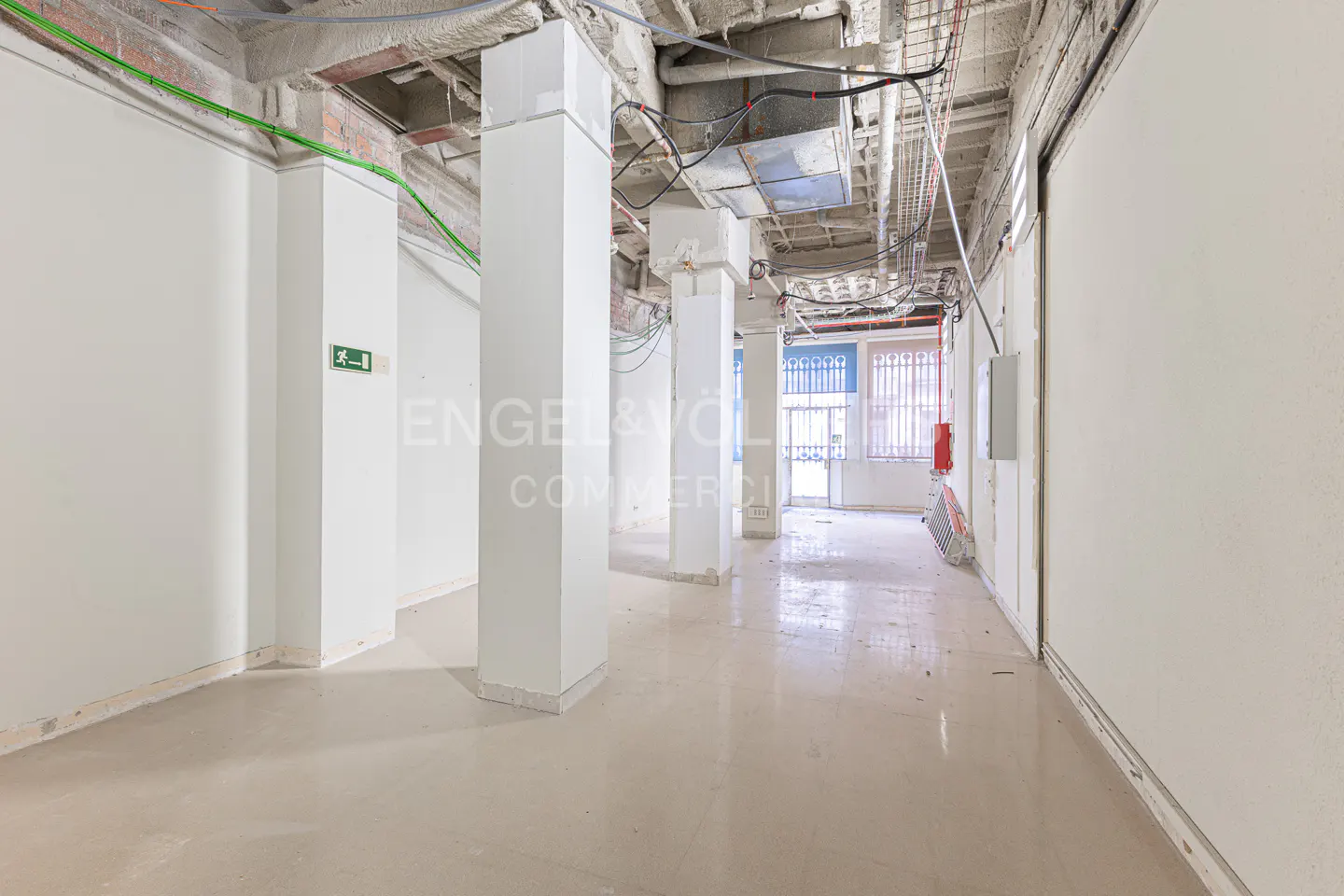 Unfinished commercial space with white walls, concrete pillars, and exposed ceiling with wiring. Exit sign visible on the left wall.
