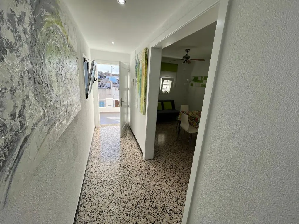 Hallway with terrazzo floor, white walls with art, and an open door to a balcony. A living room is visible through a doorway.